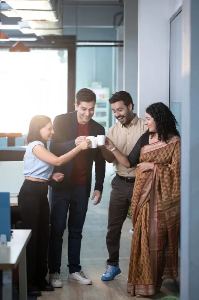 Four colleagues in a bright office toast with coffee mugs, smiling together.